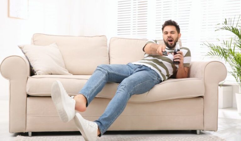 A man on a couch with a remote control, looking bored while watching TV